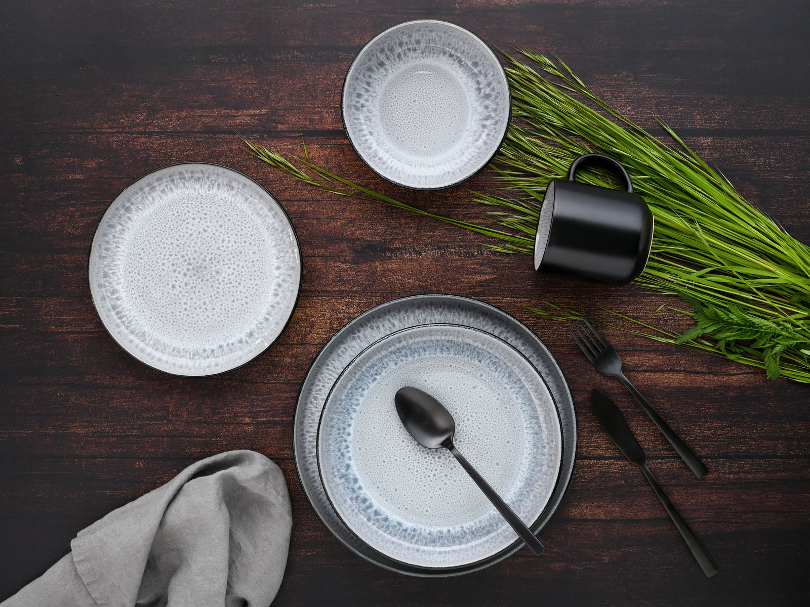 Vogelperspektive auf ein stilvolles Kombiservice-Set mit 30 Teilen in Frost-Design, bestehend aus Tellern, Schalen und einer Tasse auf einem dunklen Holztisch, dekoriert mit grünem Gras und schwarzem Besteck.