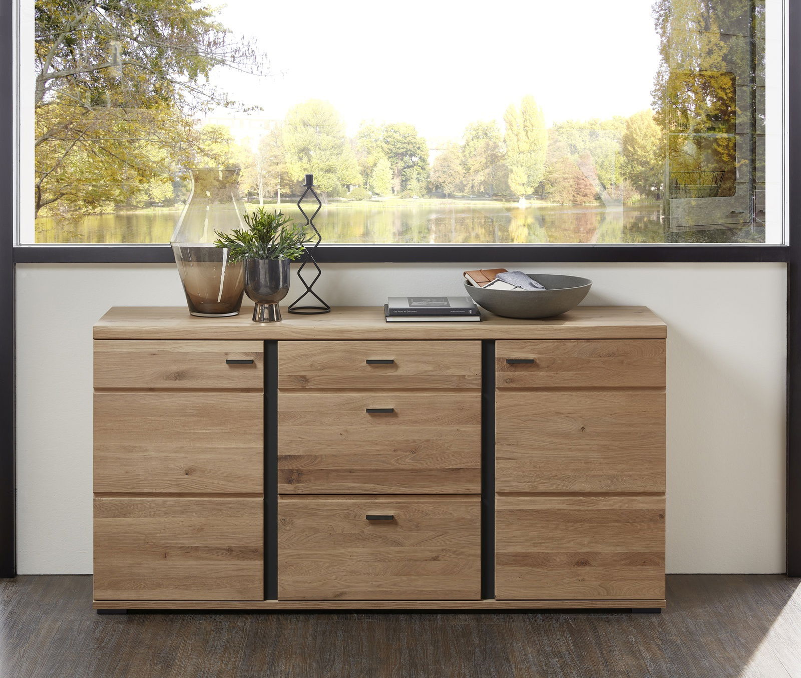 Holz-Sideboard mit mehreren Schubladen und Türen, dekoriert mit Vasen und Schalen, vor einem großen Fenster mit Blick auf einen See. Frontalansicht.