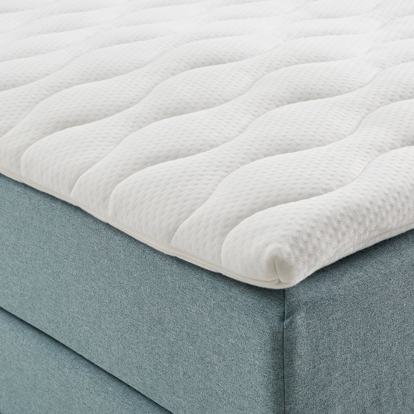 Close-up van een boxspringbed met een witte, doorgestikte topper en een lichtblauwe stoffen bekleding, gezien vanaf de zijkant