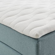 Close-up van een boxspringbed met een witte, doorgestikte topper en een lichtblauwe stoffen bekleding, gezien vanaf de zijkant