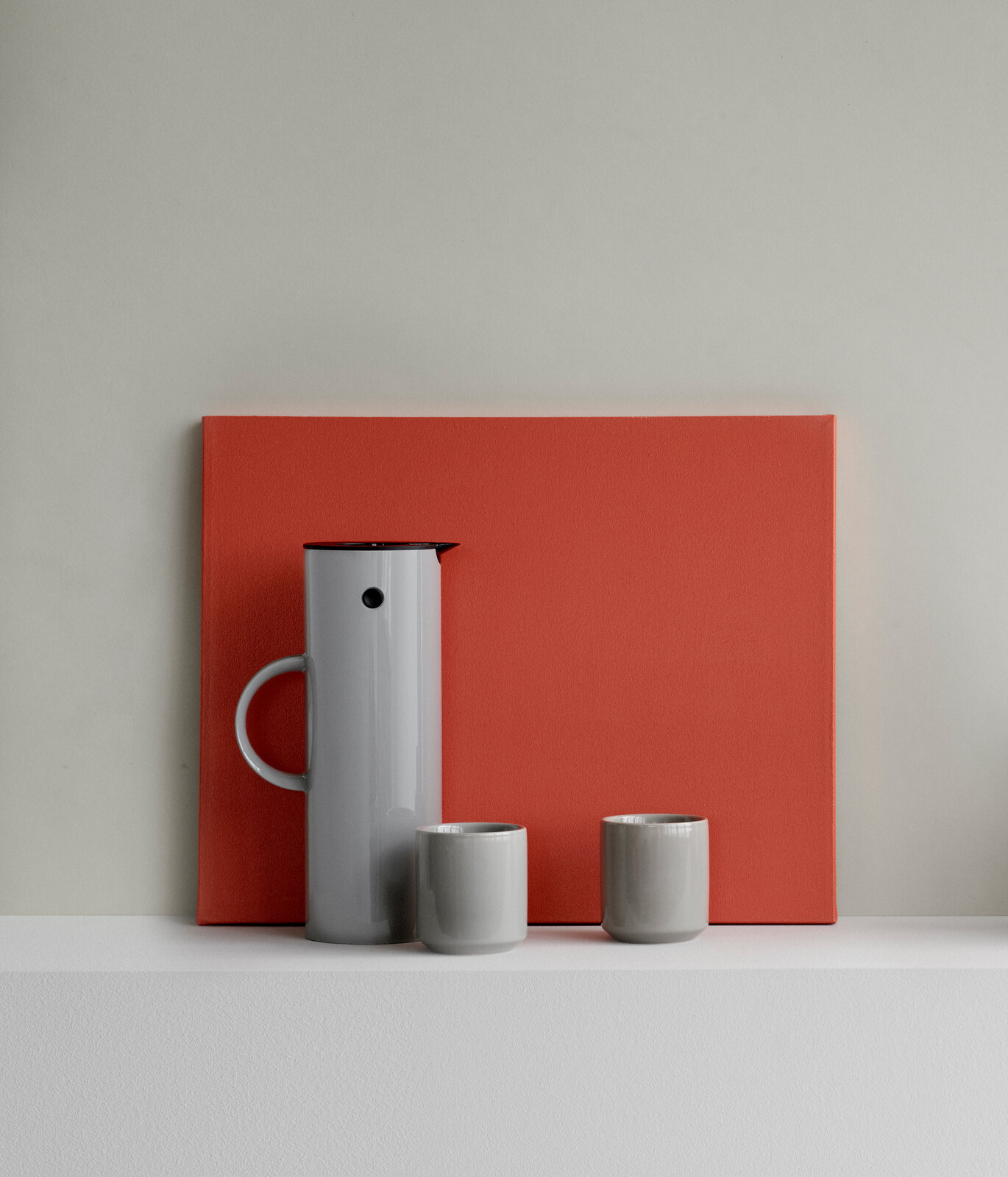 Graue Isolierkanne mit zwei passenden Tassen, frontal vor einem roten Hintergrund präsentiert.