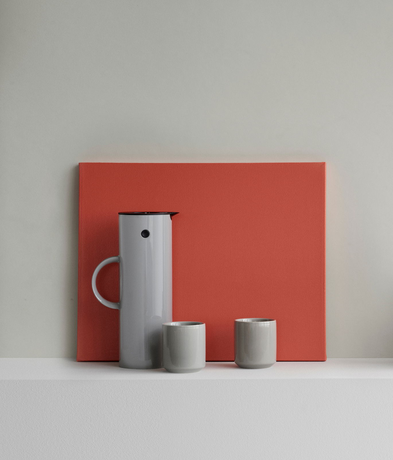 Graue Isolierkanne mit zwei passenden Tassen, frontal vor einem roten Hintergrund präsentiert.