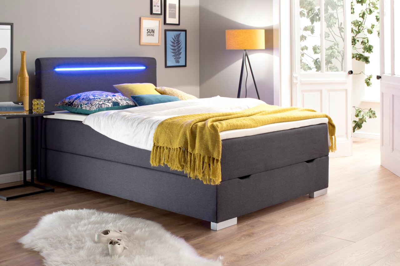 Modern boxbed met grijze stoffen bekleding, ledverlichting in het hoofdeinde en kleurrijke versieringen, zijaanzicht