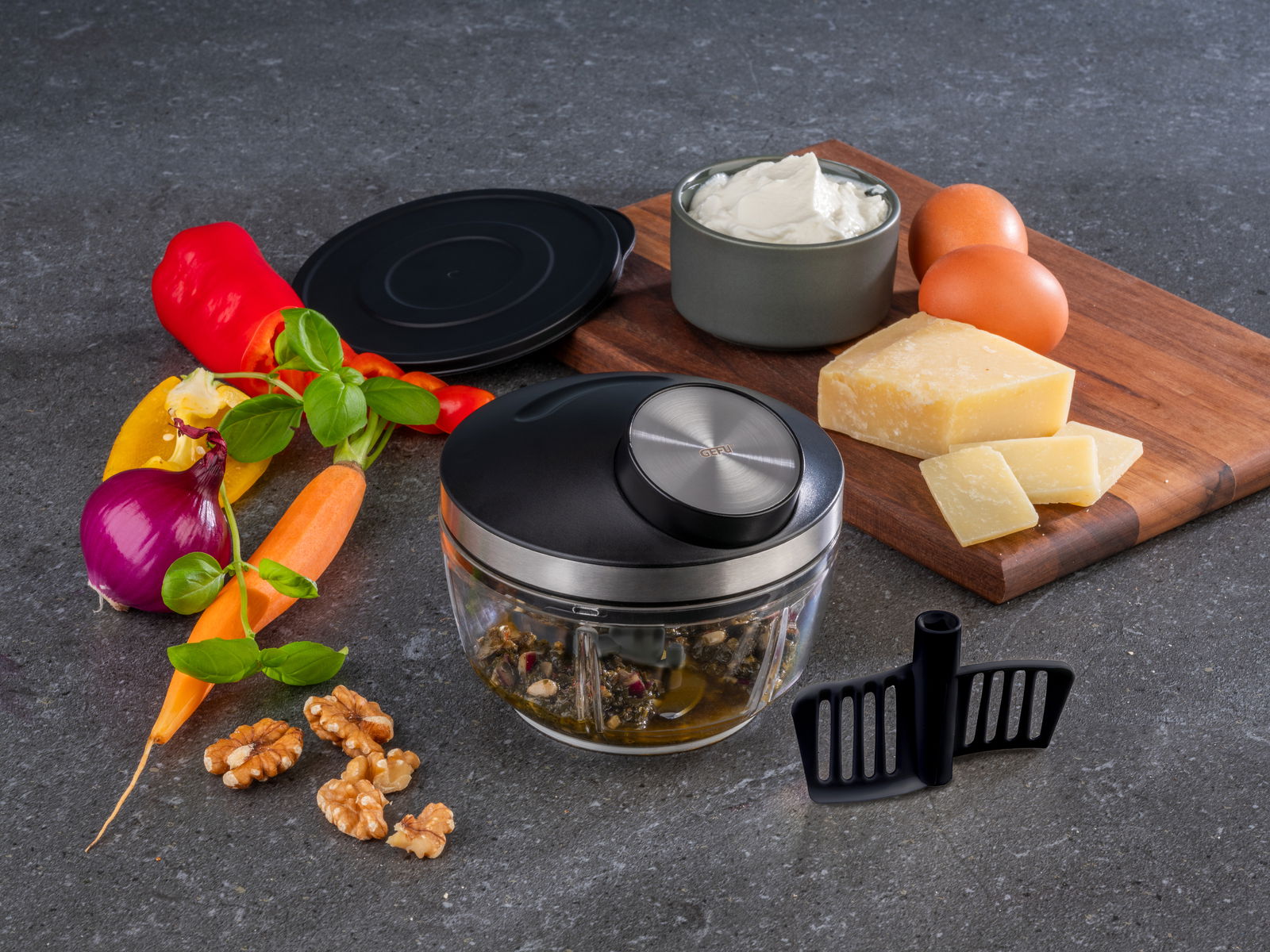 GEFU Multizerkleinerer ZIZO Multizerkleinerer ZIZO aus der Vogelperspektive, umgeben von frischen Zutaten wie Paprika, Karotten, Zwiebeln, Walnüssen, Käse und Eiern auf einer Küchenarbeitsplatte.