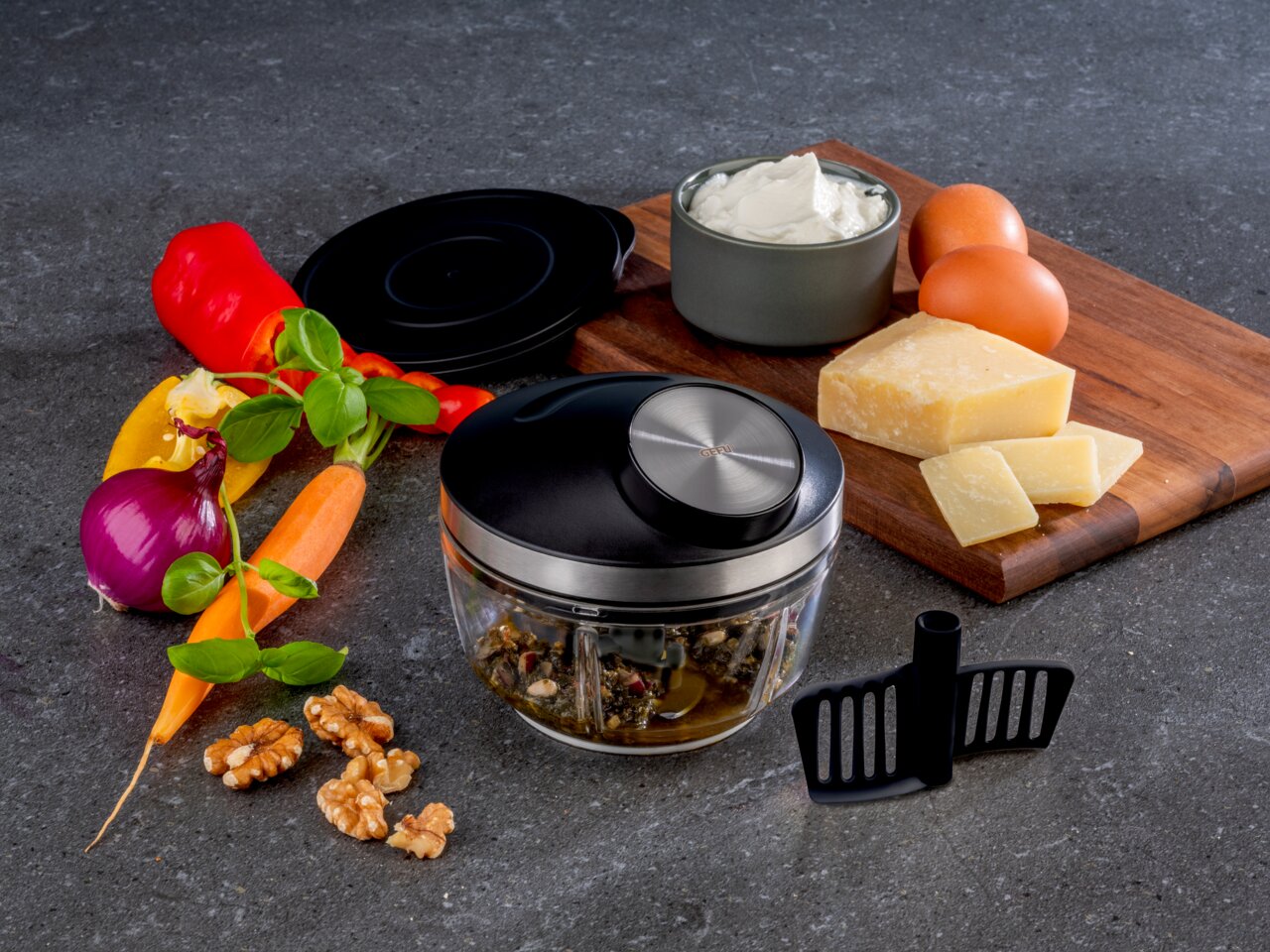 GEFU Multizerkleinerer ZIZO Multizerkleinerer ZIZO aus der Vogelperspektive, umgeben von frischen Zutaten wie Paprika, Karotten, Zwiebeln, Walnüssen, Käse und Eiern auf einer Küchenarbeitsplatte.