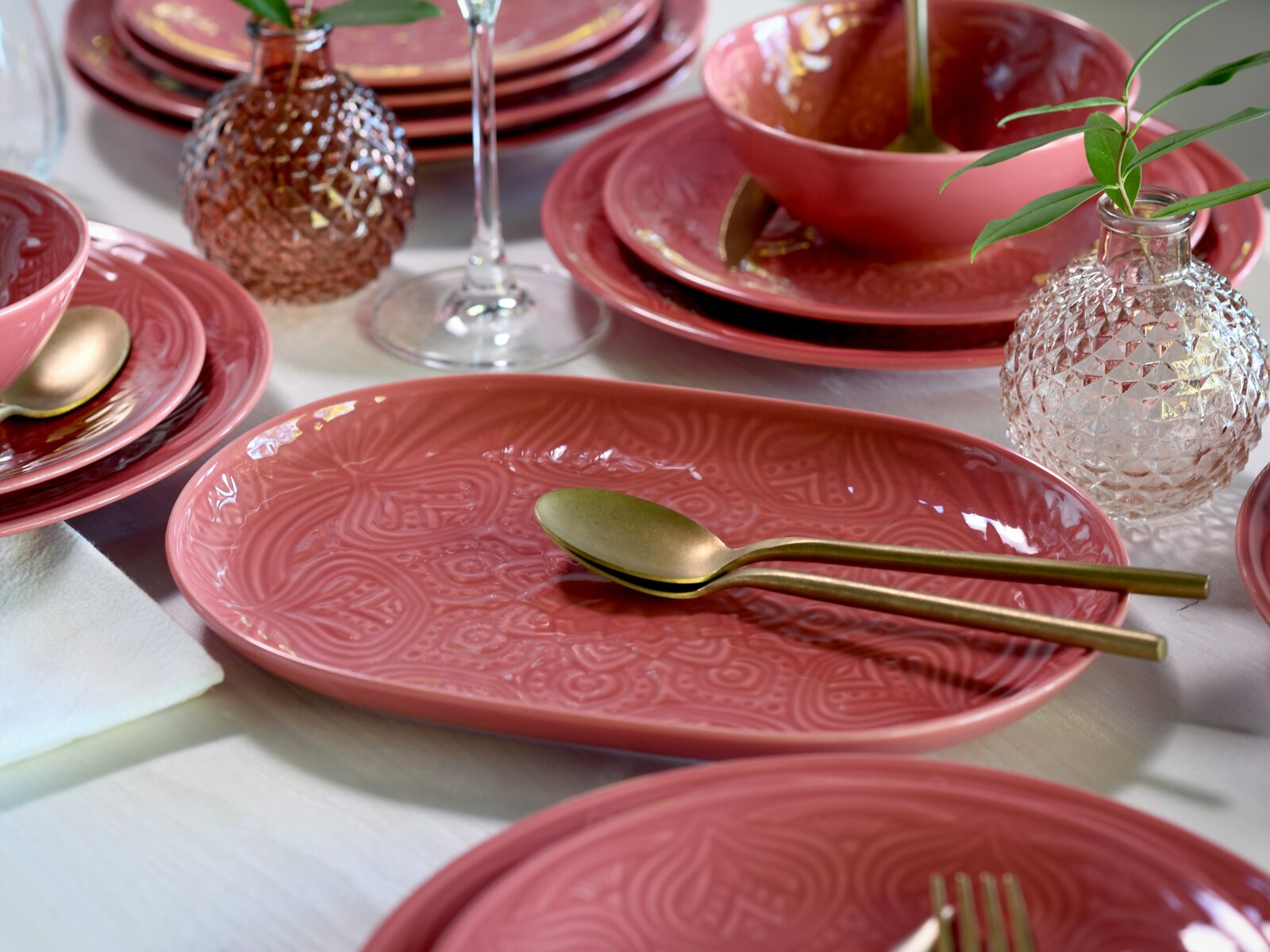 Schräg von oben fotografierte Tischdekoration mit rosafarbenen, geprägten Keramiktellern und Schalen, goldfarbenem Besteck und kleinen strukturierten Glasvasen mit grünen Zweigen auf weißer Tischdecke.