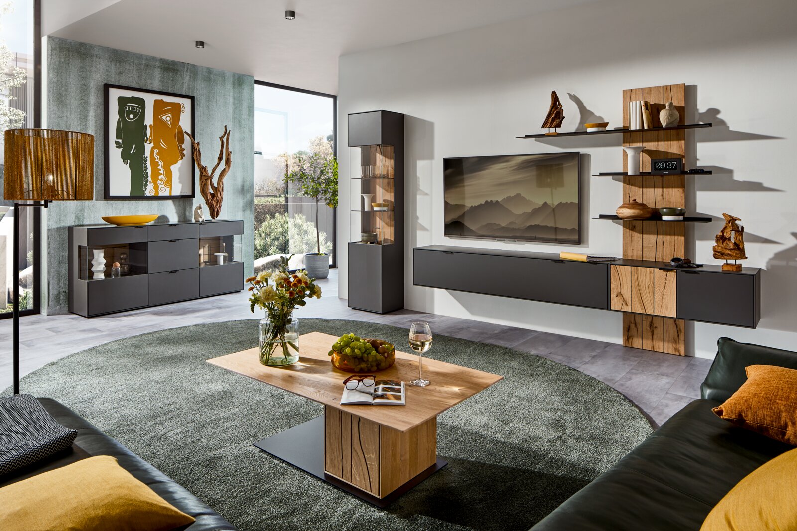 Moderne Wohnzimmer-Wandkombination in dunklen Tönen mit Holzakzenten, bestehend aus einem TV-Board, Regalen und Vitrine, aus der Perspektive eines gemütlichen Wohnzimmers mit Blick auf den Couchtisch.