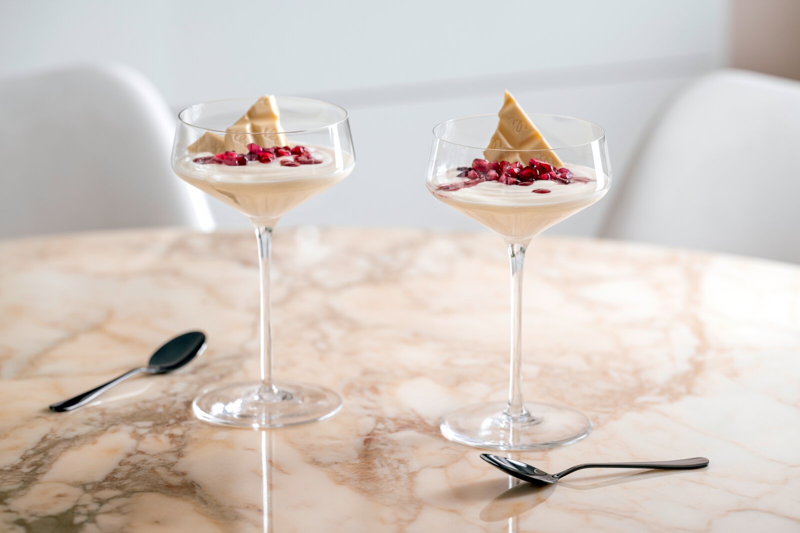 Twee elegante champagneglazen met een dessert, versierd met witte chocolade en granaatappelpitjes, gezien vanuit een zijaanzicht op een marmeren tafel.