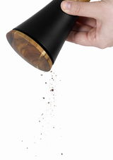 Hand hält eine schwarze Pfeffer- oder Salzmühle mit Holzboden, aus der Gewürze herausrieseln. Perspektive von der Seite.