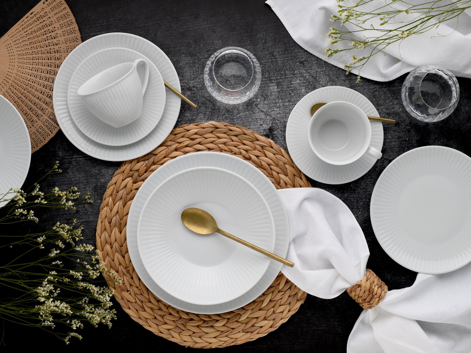 Draufsicht auf ein elegantes weißes Porzellan-Geschirrset mit gerillten Tellern und Tassen, goldfarbenem Besteck, geflochtenen Platzsets, Glas-Teelichthaltern und weißer Serviette auf dunklem Tisch.