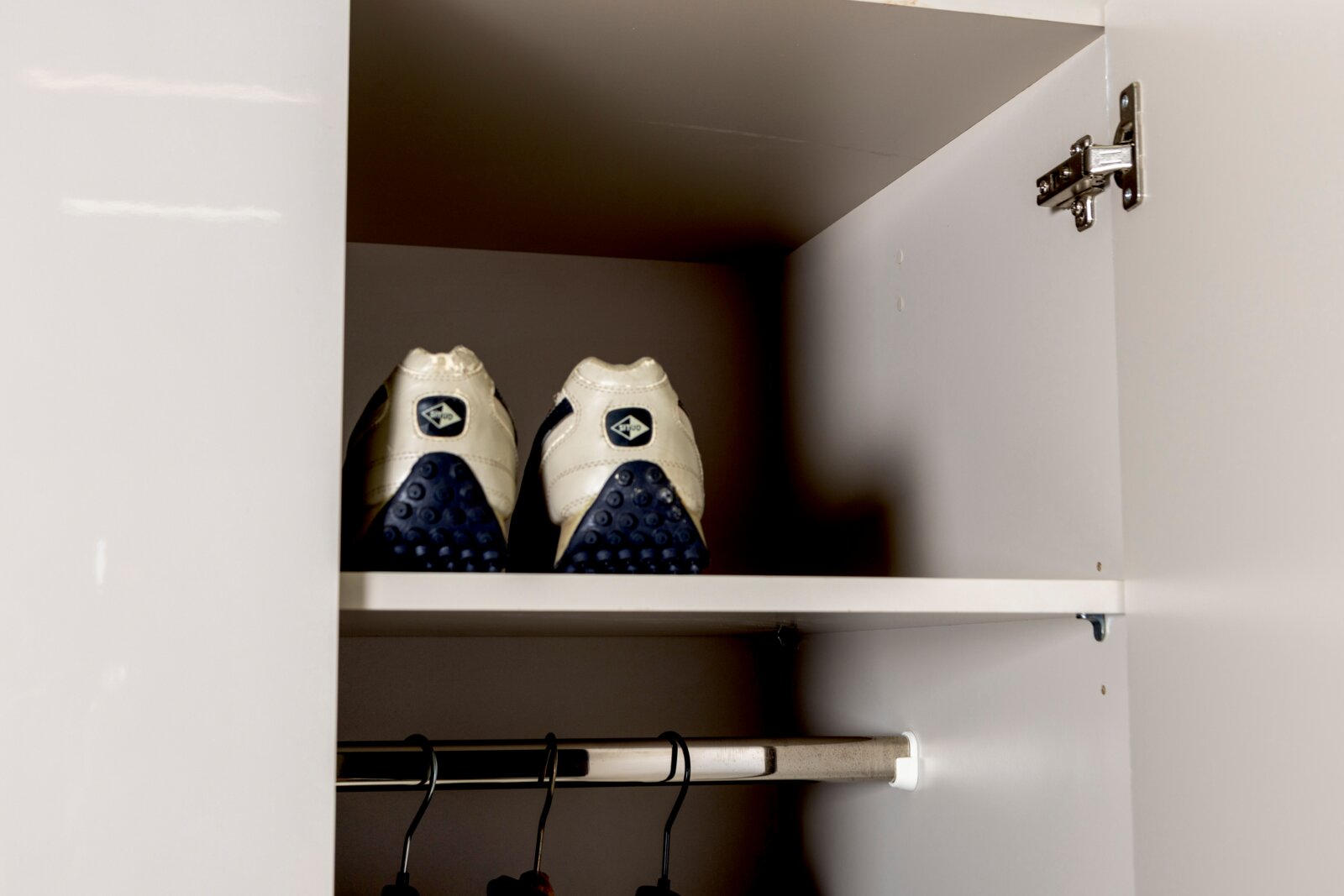 Offener weißer Kleiderschrank mit Einlegeboden und Kleiderstange; oben stehen zwei weiße Sportschuhe mit blauer Sohle, Frontansicht aus dem Schrankinneren