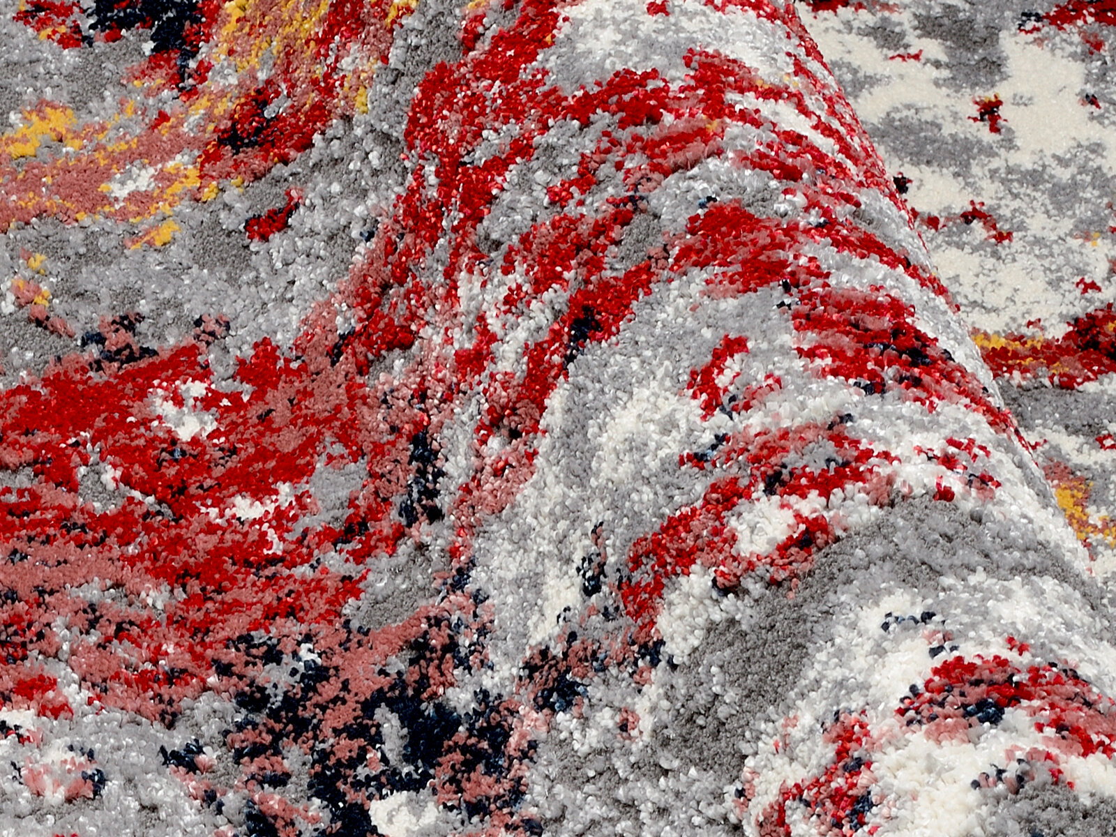 Nahaufnahme eines Designer-Teppichs mit abstraktem Muster in Rot-, Grau- und Gelbtönen, aus seitlicher Perspektive aufgenommen.