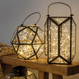 Zwei geometrische Laternen mit LED-Lichterketten, eine in Dodekaederform und eine in zylindrischer Form, auf einem Holztisch aus seitlicher Perspektive.