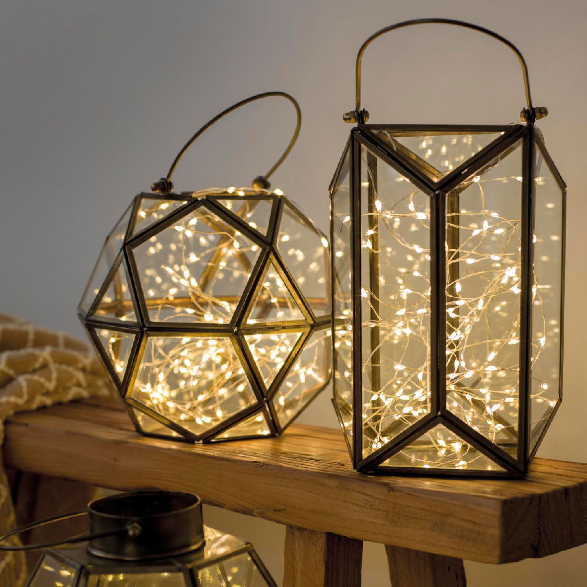 Zwei geometrische Laternen mit LED-Lichterketten, eine in Dodekaederform und eine in zylindrischer Form, auf einem Holztisch aus seitlicher Perspektive.