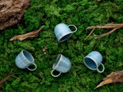 Drei hellblaue, glasierte Keramik-Kaffeetassen mit Henkel liegen dekorativ auf grünem Moos zwischen Holzstücken, Draufsicht von oben.