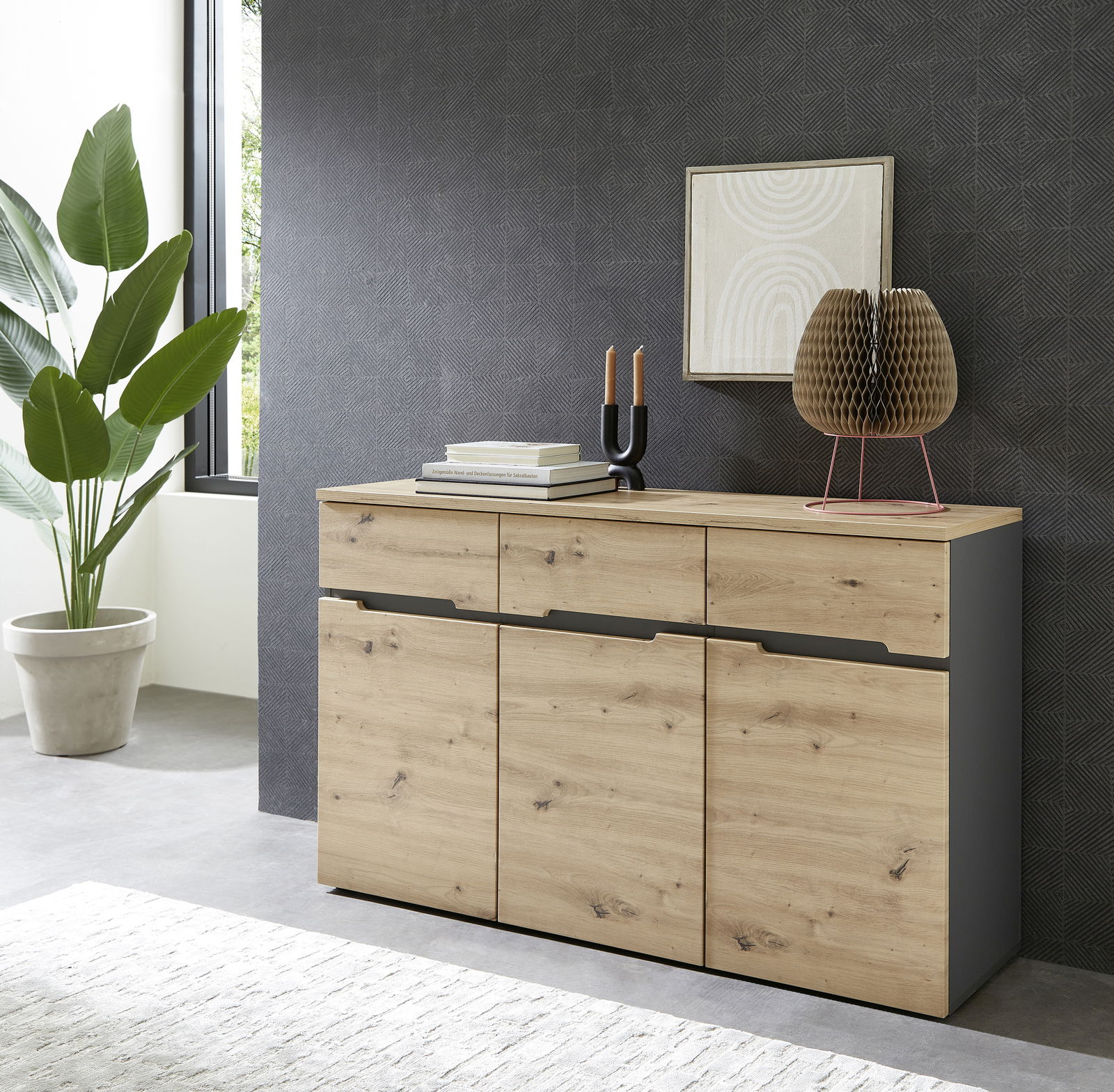 Modernes Sideboard aus Holz mit drei Schubladen und drei Türen, dekoriert mit Büchern, Kerzen und einer Skulptur, seitliche Perspektive.