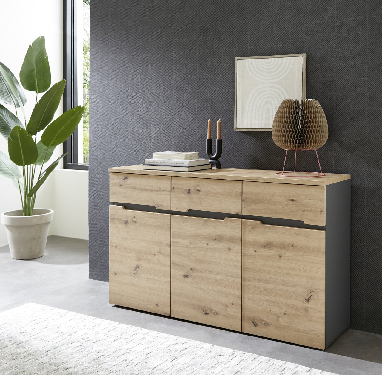Voleo Sideboard DETROIT Modernes Sideboard aus Holz mit drei Schubladen und drei Türen, dekoriert mit Büchern, Kerzen und einer Skulptur, seitliche Perspektive.