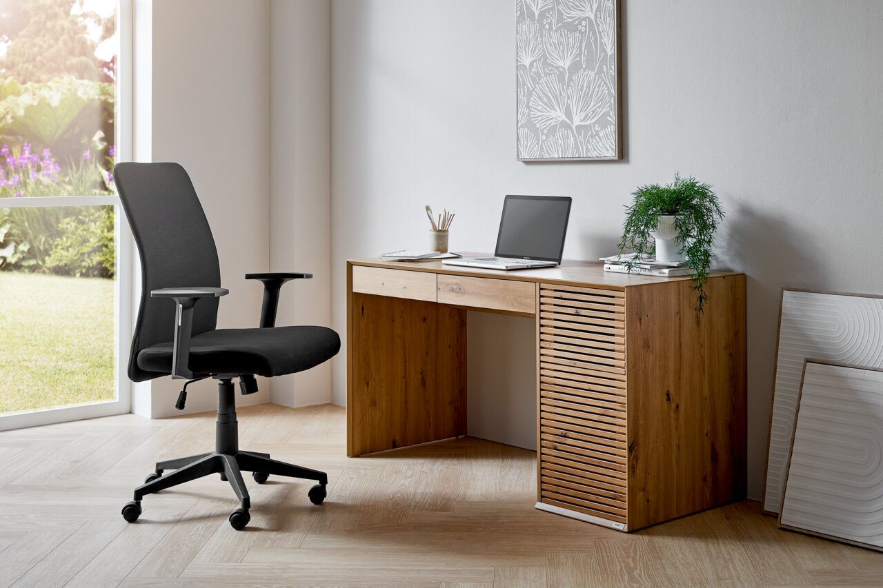 Moderne zwarte draaistoel met armleuningen in een lichte ruimte, naast een houten tafel met laptop, gezien vanuit de zijkant.