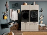 Modern eingerichteter Hauswirtschaftsraum mit beige-weißer Schrankwand und integrierter schwarzer Waschmaschine und Trockner, daneben Waschbecken, Wäsche auf Kleiderstange und geflochtene Körbe, frontal aus Augenhöhe fotografiert.