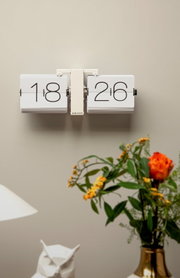 Wanduhr mit Klappzahlen in modernem Design, frontal an der Wand montiert, umgeben von Dekorationen wie einer Vase mit Blumen.