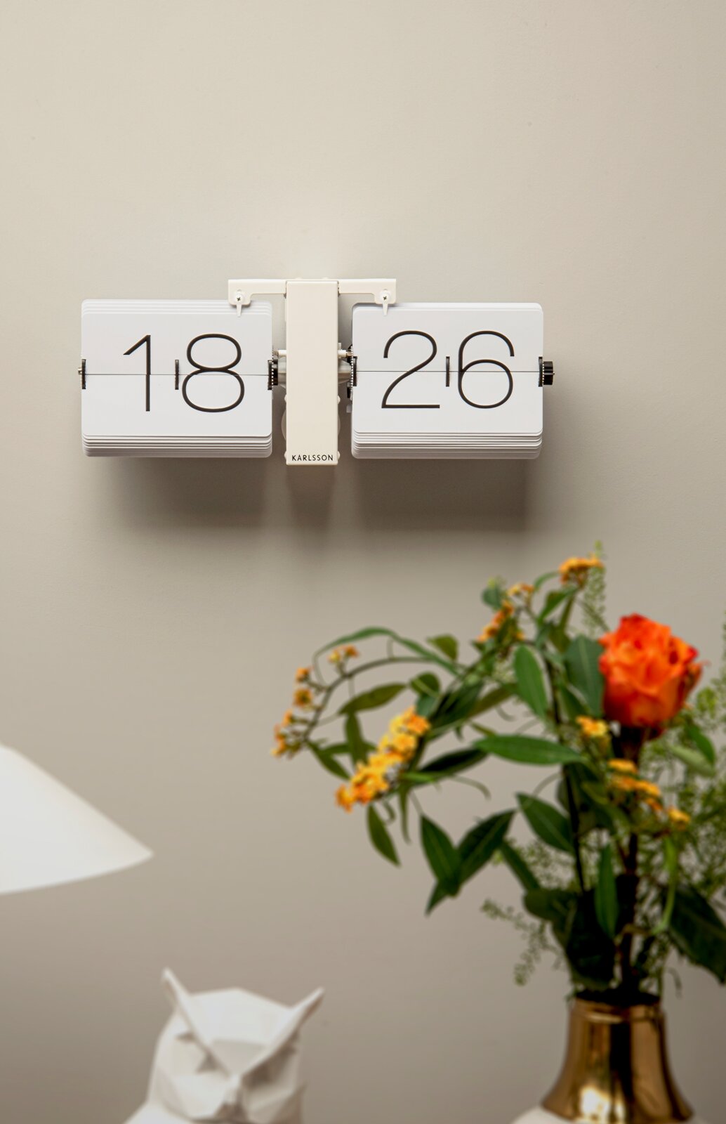 Wanduhr mit Klappzahlen in modernem Design, frontal an der Wand montiert, umgeben von Dekorationen wie einer Vase mit Blumen.