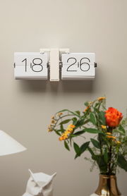 Wanduhr mit Klappzahlen in modernem Design, frontal an der Wand montiert, umgeben von Dekorationen wie einer Vase mit Blumen.