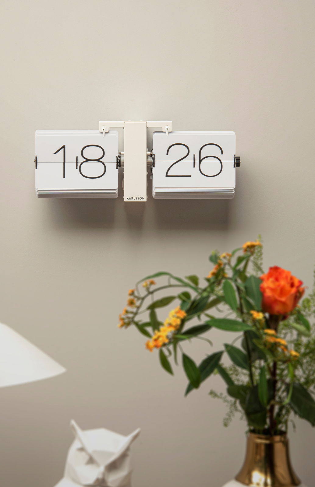 Wanduhr mit Klappzahlen in modernem Design, frontal an der Wand montiert, umgeben von Dekorationen wie einer Vase mit Blumen.