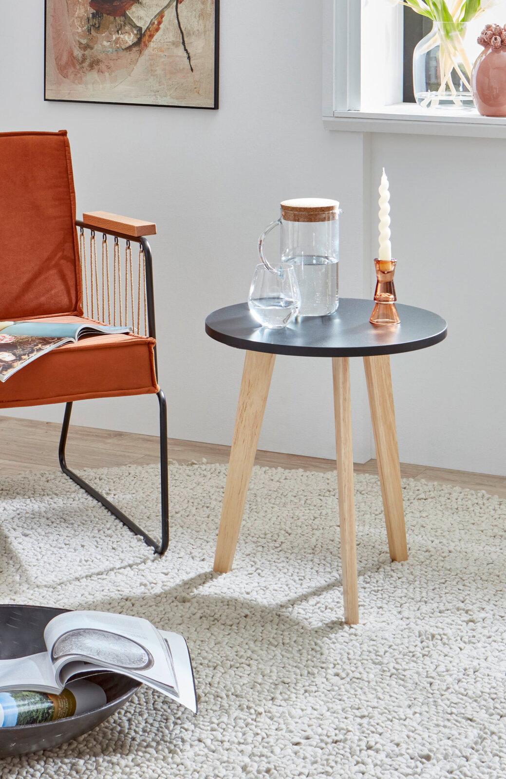 Runder Beistelltisch mit schwarzer Tischplatte und hellen Holzbeinen im Wohnzimmer-Setting, seitliche Perspektive mit Stuhl und Deko auf dem Tisch