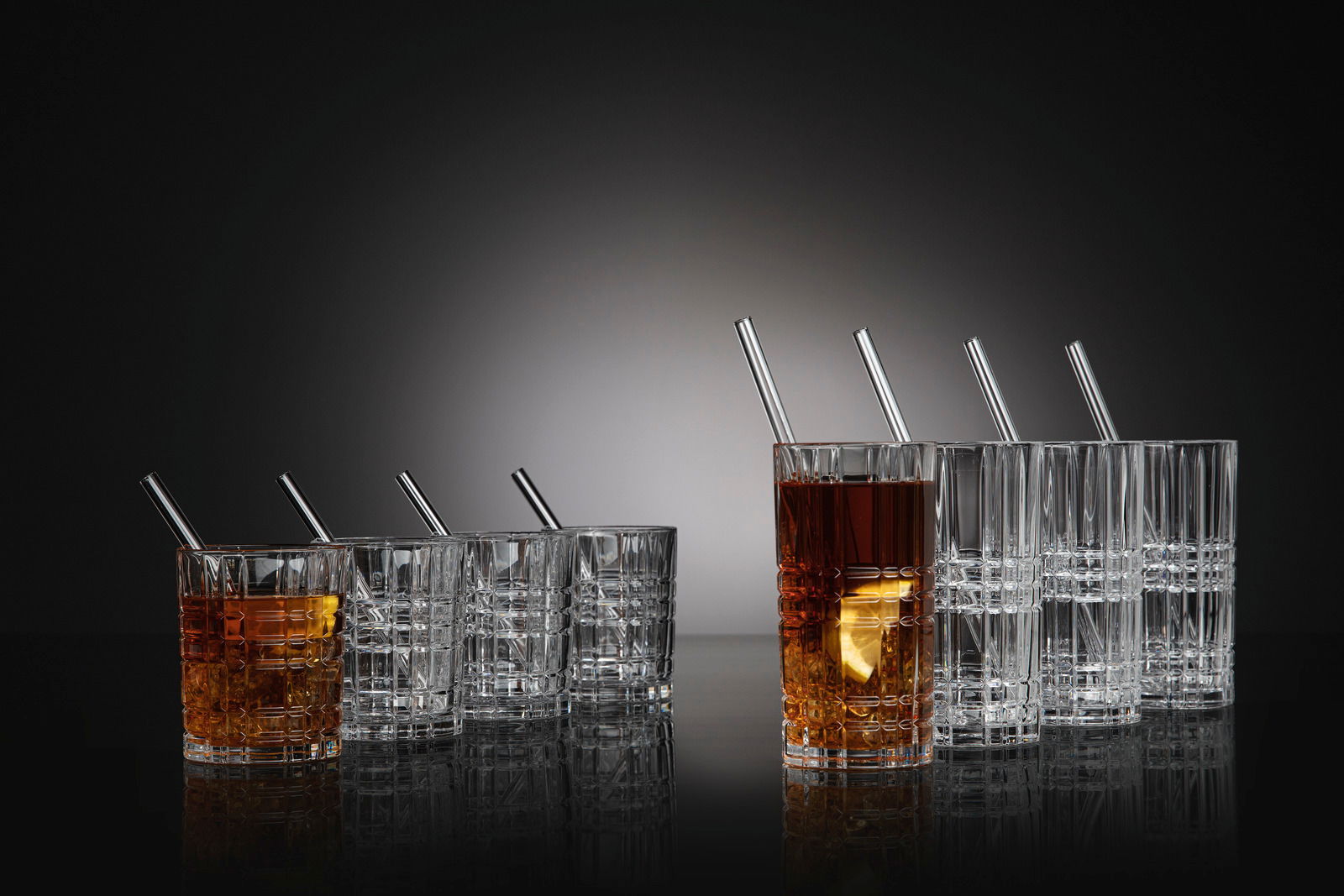 Set aus acht Gläsern mit Strohhalmen, teilweise gefüllt mit Getränken, auf einer reflektierenden Oberfläche, seitliche Perspektive.