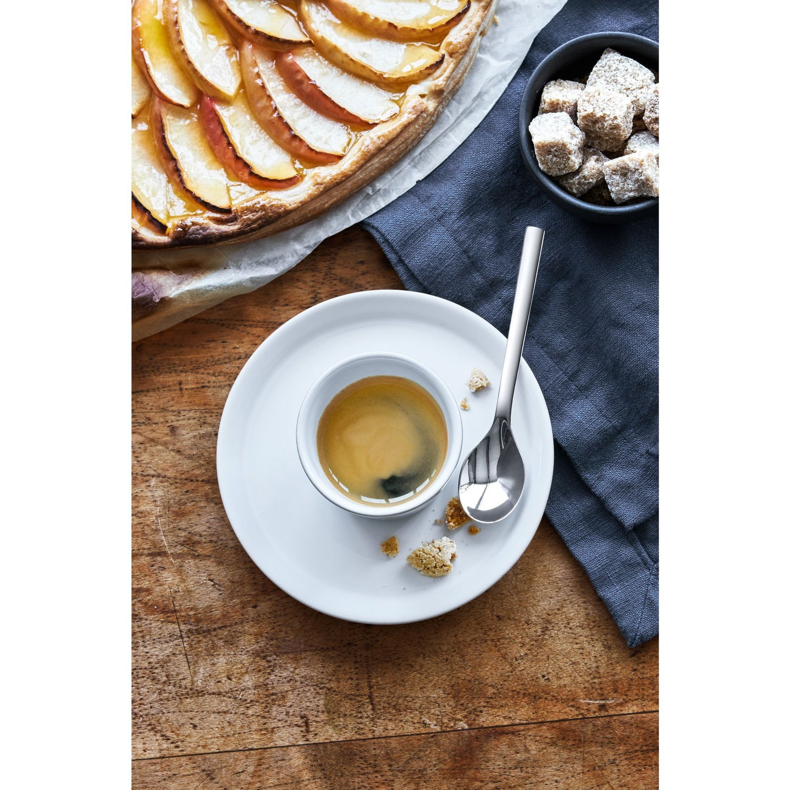 Espressolöffel auf einer Untertasse mit Espresso, von oben fotografiert, neben einem Apfelkuchen und Zuckerwürfeln.