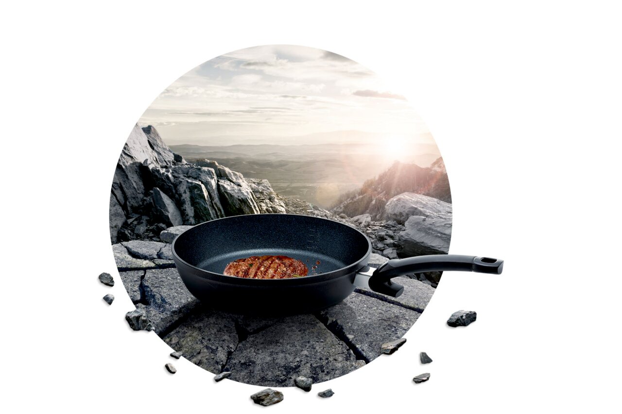 Pfanne Adamant Comfort mit gebratenem Steak, auf einem Felsen in einer Berglandschaft platziert, seitliche Perspektive.