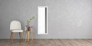 Rechteckiger Wandspiegel mit weißem Rahmen, vertikal an einer grauen Wand montiert, neben einem Stuhl und einem kleinen Tisch mit Dekorationen, aus seitlicher Perspektive.
