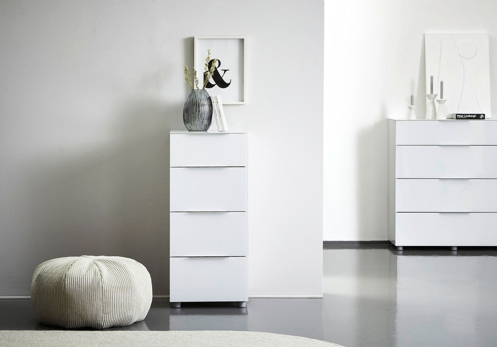 Voleo Hochkommode MALIN Weiße Hochkommode mit vier Schubladen, dekoriert mit einer Vase und Büchern, in einem modernen, minimalistischen Raum. Perspektive von vorne.