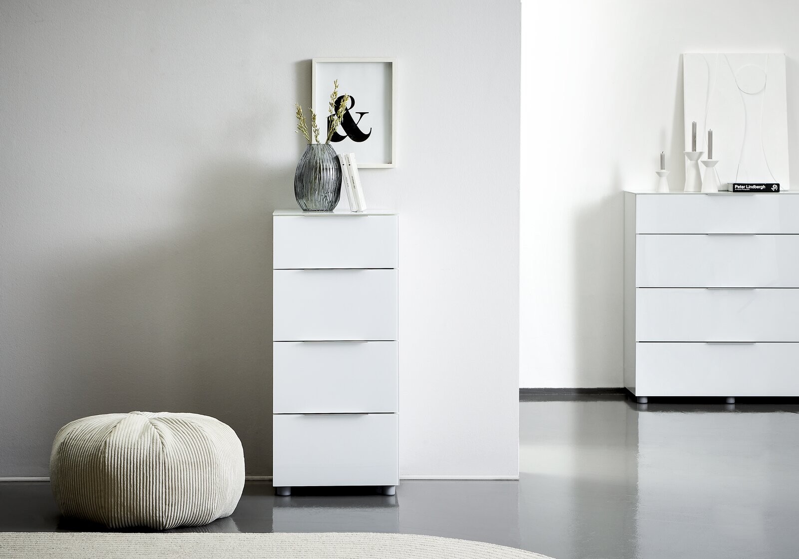 Voleo Hochkommode MALIN Weiße Hochkommode mit vier Schubladen, dekoriert mit einer Vase und Büchern, in einem modernen, minimalistischen Raum. Perspektive von vorne.