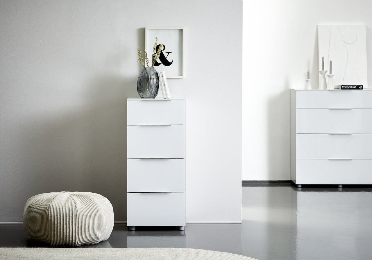 Weiße Hochkommode mit vier Schubladen, dekoriert mit einer Vase und Büchern, in einem modernen, minimalistischen Raum. Perspektive von vorne.