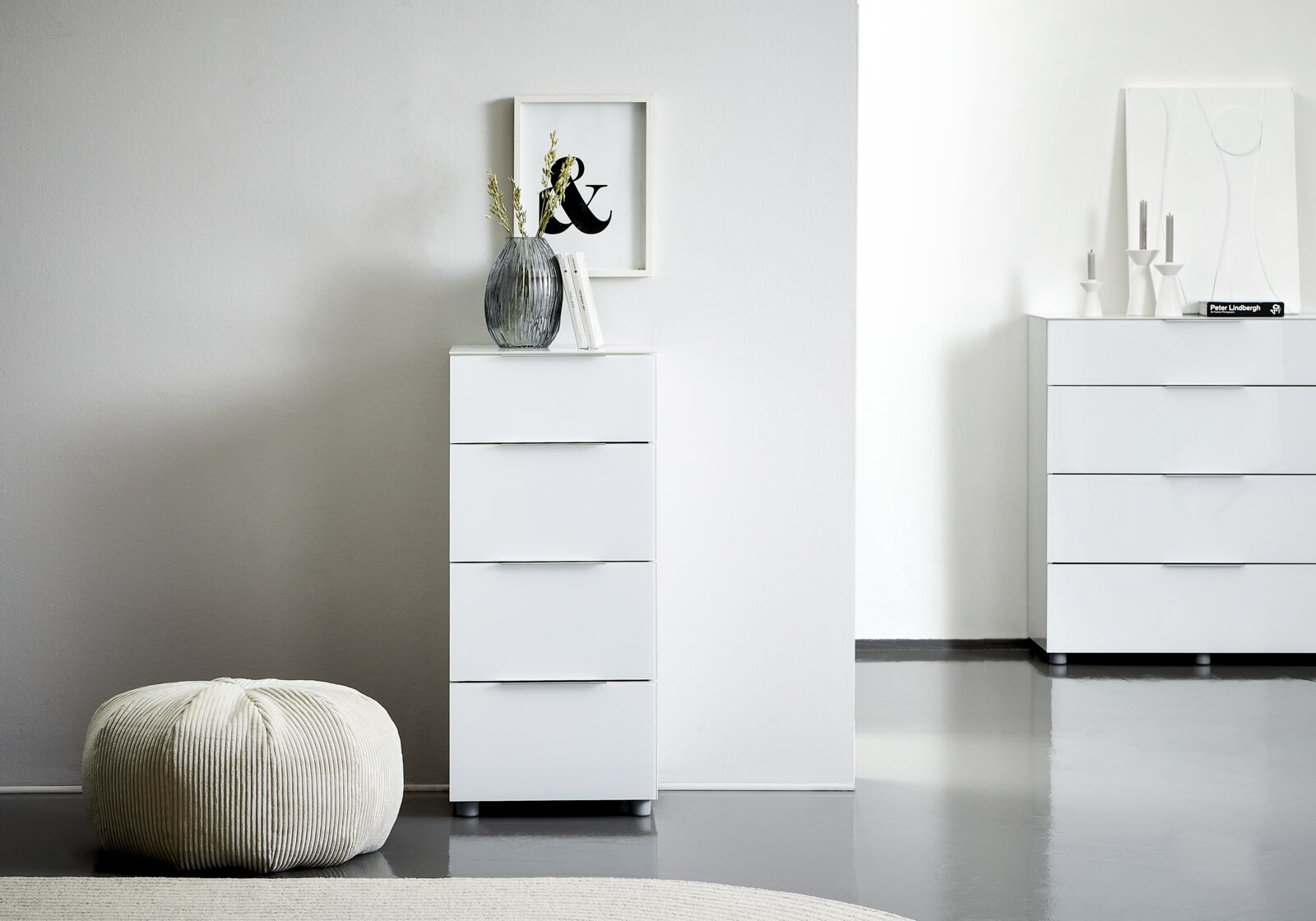Weiße Hochkommode mit vier Schubladen, dekoriert mit einer Vase und Büchern, in einem modernen, minimalistischen Raum. Perspektive von vorne.