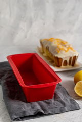 Rote Silikon-Kastenform in Nahaufnahme, auf einem grauen Tuch liegend, mit einem Kuchen im Hintergrund.