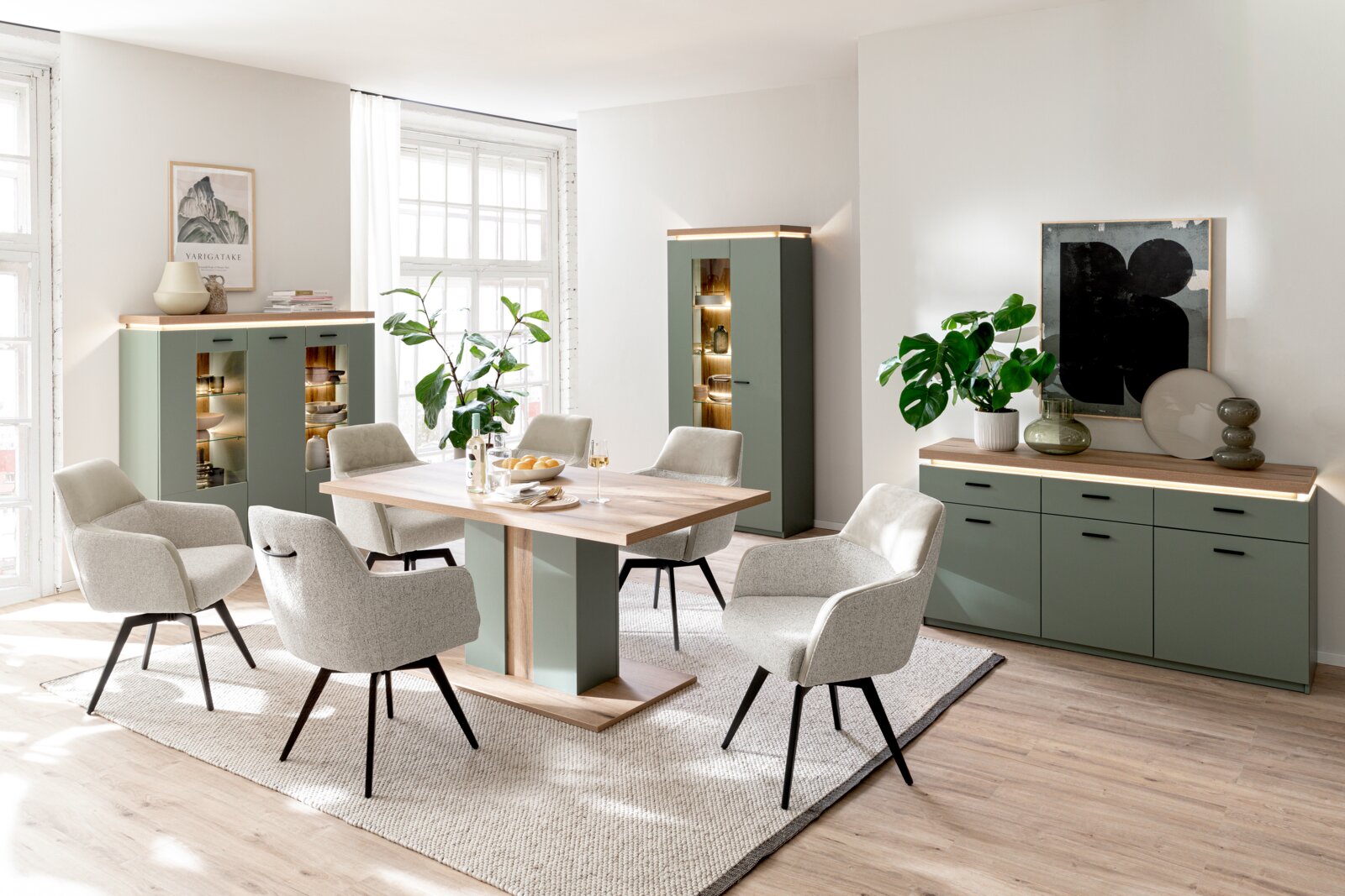 Helles Esszimmer-Set mit rechteckigem Esstisch in Holzoptik und grünem Mittelfuß, umgeben von sechs gepolsterten Stühlen, dazu grüne Sideboards und Vitrinenschrank mit LED-Beleuchtung Perspektive: frontale Weitwinkel-Ansicht auf die komplette Raumgruppe.