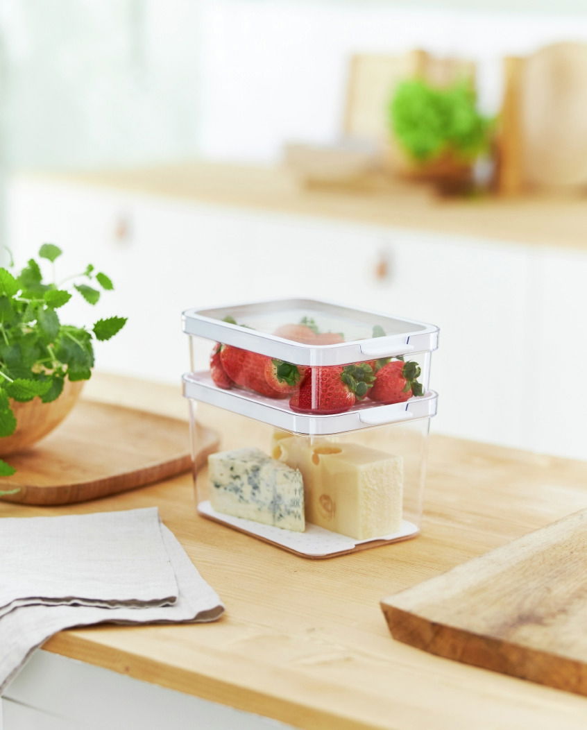 Zwei transparente Kühlschrankorganizer-Boxen, übereinander gestapelt, gefüllt mit Erdbeeren und Käse, auf einer Küchenarbeitsplatte aus Holz, seitliche Perspektive.