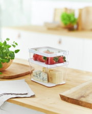 Zwei transparente Kühlschrankorganizer-Boxen, übereinander gestapelt, gefüllt mit Erdbeeren und Käse, auf einer Küchenarbeitsplatte aus Holz, seitliche Perspektive.