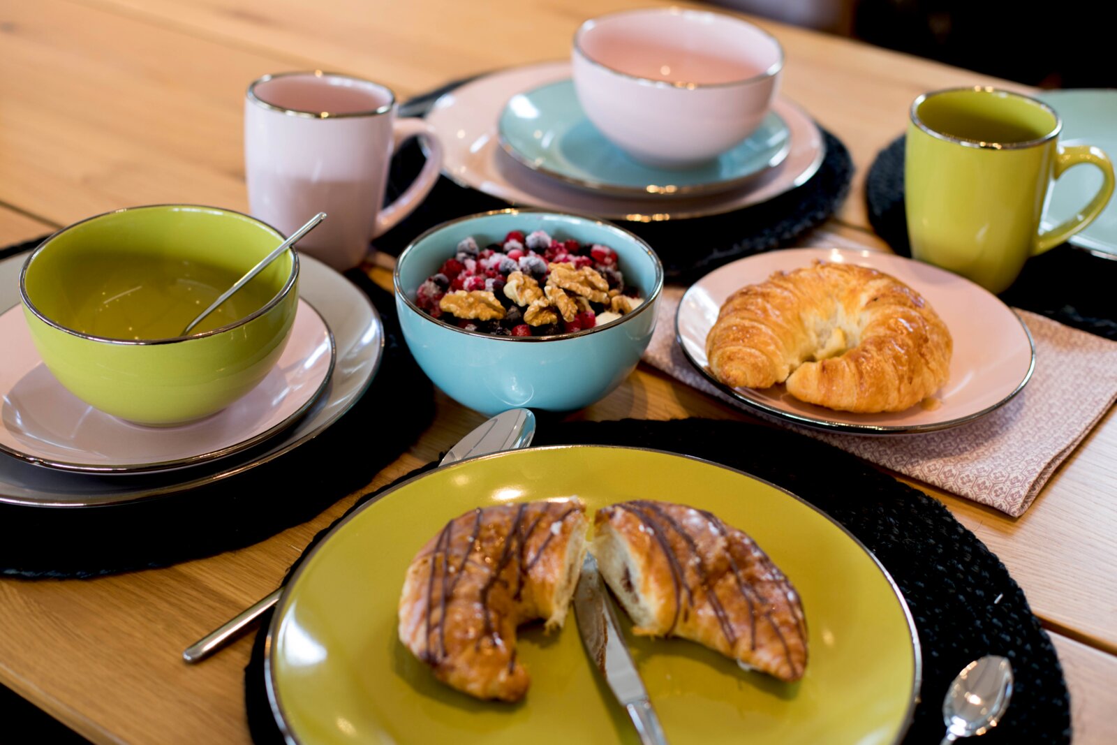 Schick gedeckter Frühstückstisch mit buntem Geschirr in Grün, Blau und Rosa, Müsli mit Beeren und Walnüssen sowie Croissants auf Holztisch, fotografiert aus leicht schräger Draufsicht.