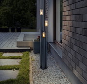 Moderne Außenleuchte mit drei vertikalen Leuchtkörpern, installiert neben einer Hauswand auf einem Kiesbett, mit Blick auf eine Terrasse und Garten.