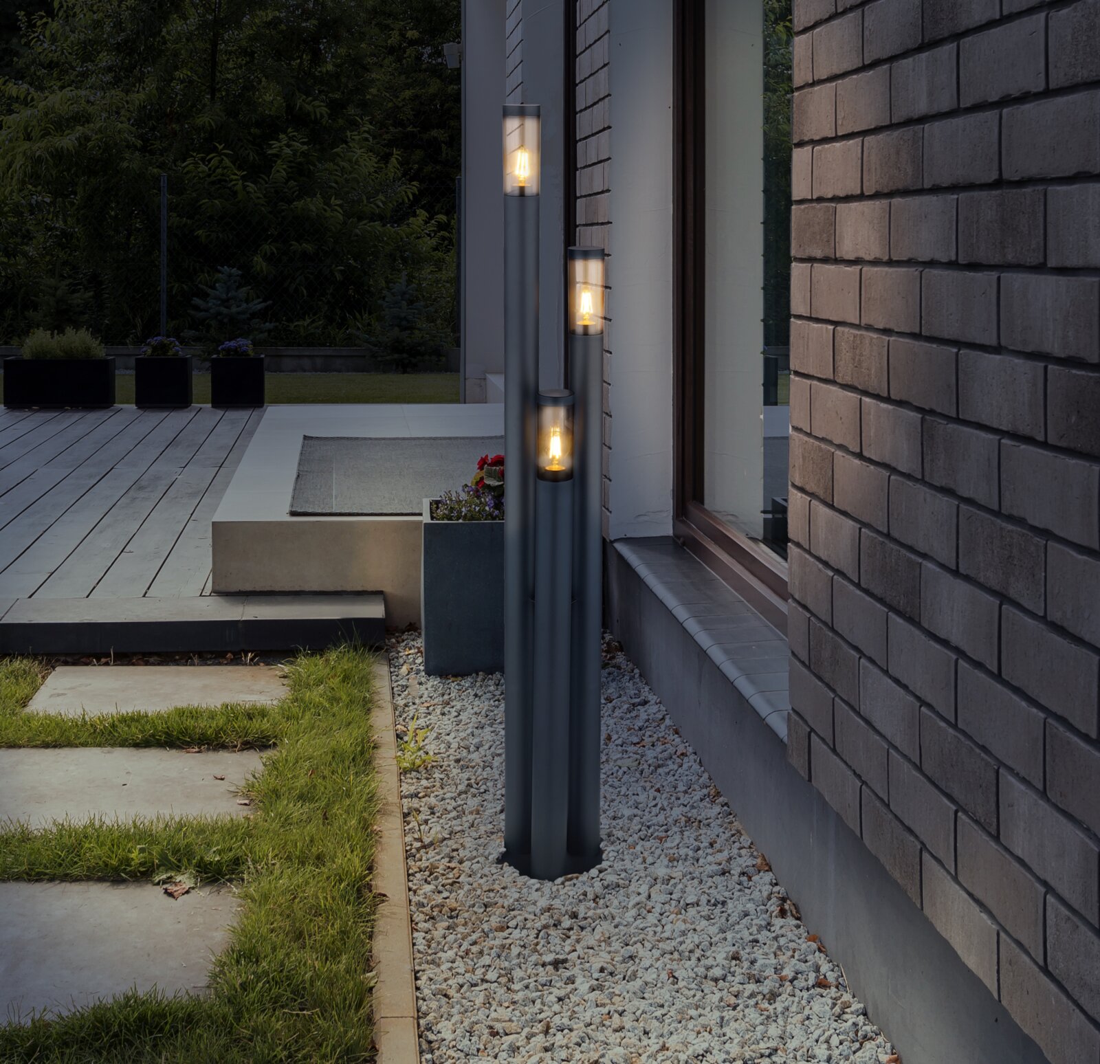 Moderne Außenleuchte mit drei vertikalen Leuchtkörpern, installiert neben einer Hauswand auf einem Kiesbett, mit Blick auf eine Terrasse und Garten.