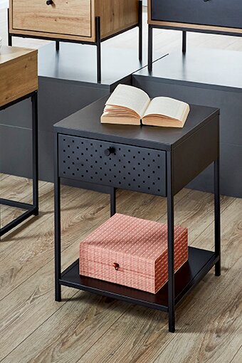 Moderner schwarzer Beistelltisch aus Metall mit perforierter Schubladenfront, Ablageboden und geöffnetem Buch auf der Tischplatte, frontal in leicht erhöhter Perspektive fotografiert
