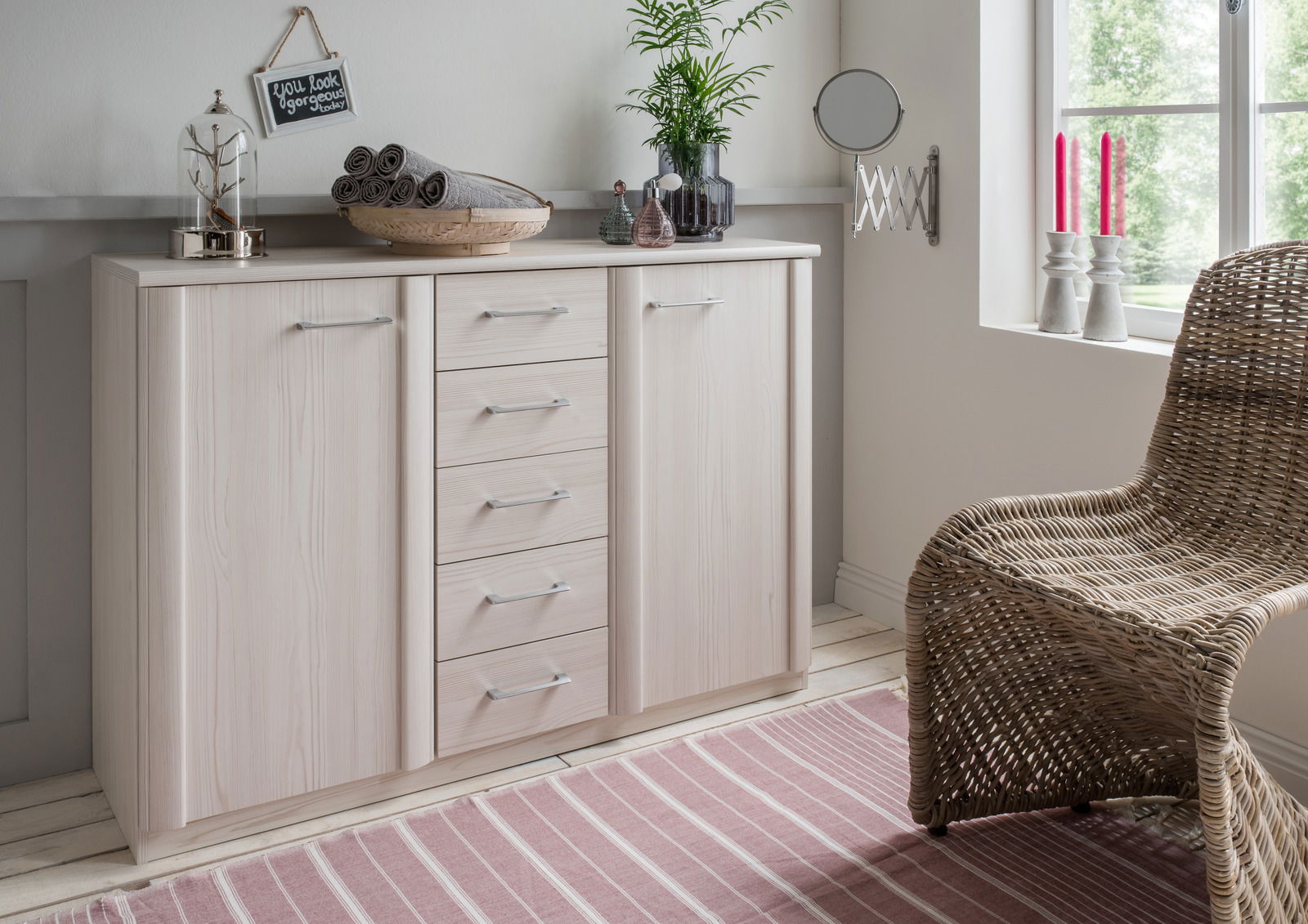 Helle Kombikommode mit mehreren Schubladen und Türen, dekoriert mit Pflanzen und Accessoires, in einem gemütlichen Raum mit Fenster und Korbstuhl, aus seitlicher Perspektive.