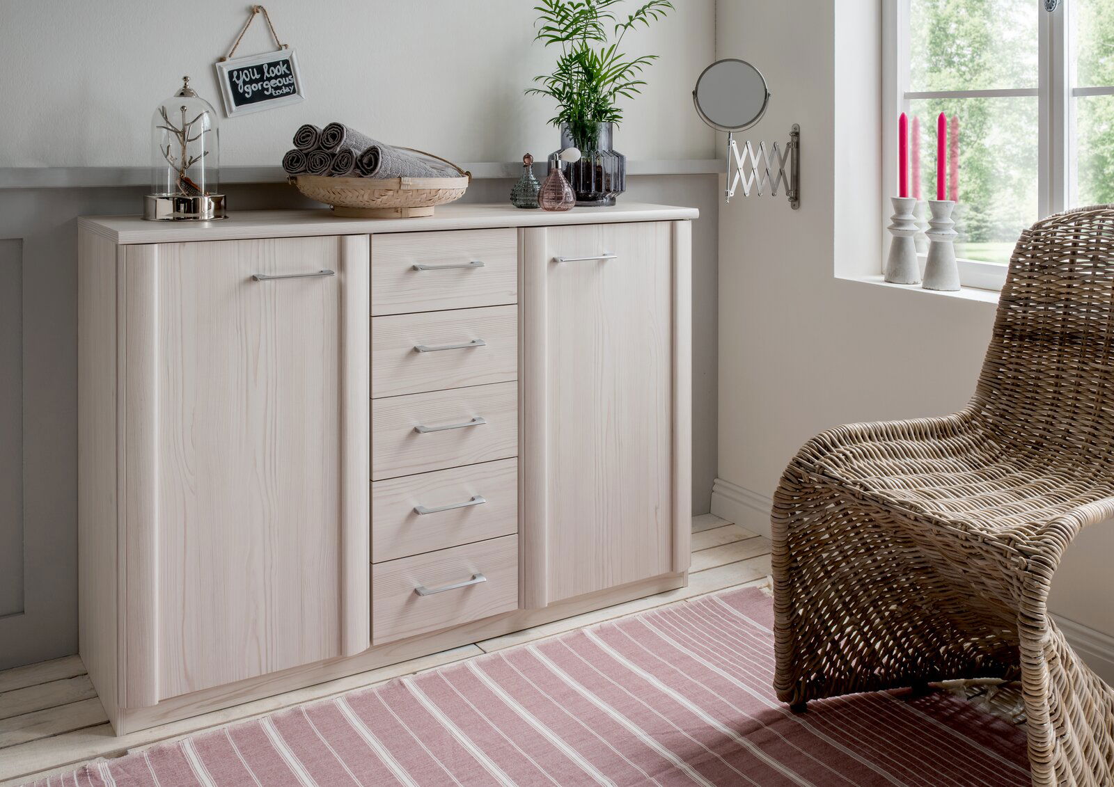 Venda Kombikommode LUXOR Helle Kombikommode mit mehreren Schubladen und Türen, dekoriert mit Pflanzen und Accessoires, in einem gemütlichen Raum mit Fenster und Korbstuhl, aus seitlicher Perspektive.