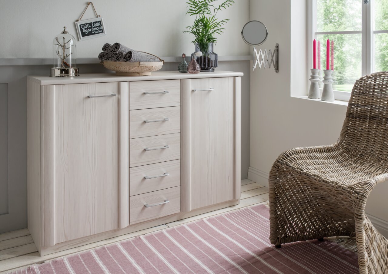 Venda Kombikommode LUXOR Helle Kombikommode mit mehreren Schubladen und Türen, dekoriert mit Pflanzen und Accessoires, in einem gemütlichen Raum mit Fenster und Korbstuhl, aus seitlicher Perspektive.