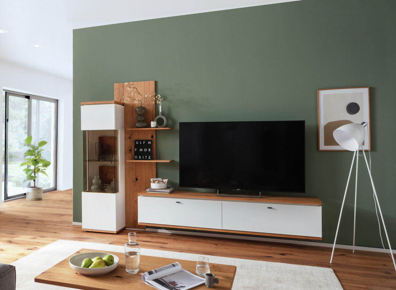 Novel Wandkombination MATADI Moderne Wandkombination MATADI in einem stilvollen Wohnzimmer mit grüner Wand, bestehend aus einem TV-Schrank und einem Vitrinenschrank, aus Holz und weißen Elementen, seitliche Perspektive.