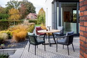 Moderner Designsessel auf einer Terrasse, umgeben von einem runden Holztisch und weiteren Sesseln, mit Blick auf einen Garten im Herbst.