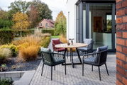 Moderner Designsessel auf einer Terrasse, umgeben von einem runden Holztisch und weiteren Sesseln, mit Blick auf einen Garten im Herbst.